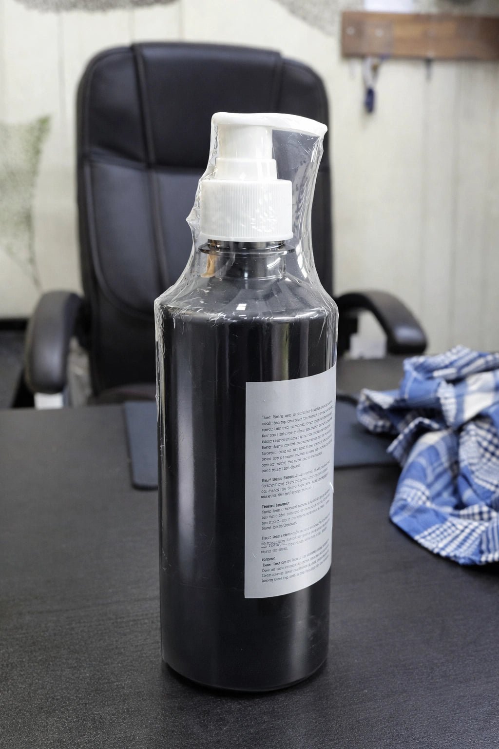 Black spray bottle with white pump dispenser placed on office desk beside folded blue plaid cloth and black chair